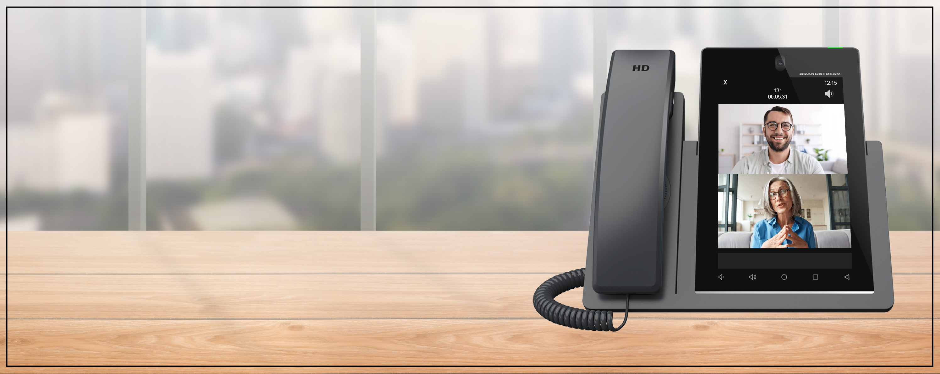 Grandstream video desk phone showing a two-way video call, on a wooden desk by an office window